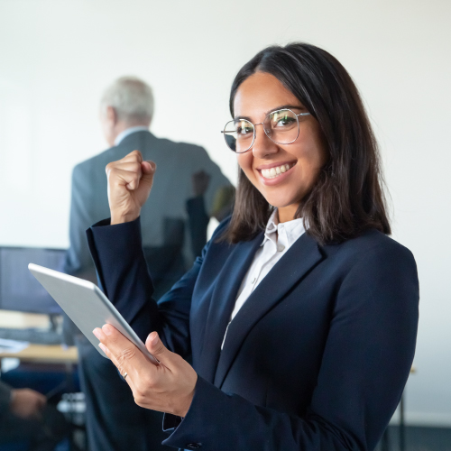 Una mujer vestida formalmente sostiene una tableta en una mano mientras levanta el puño en un gesto de celebración. Detrás de ella, un hombre mayor, también vestido formalmente, observa una computadora. La escena tiene lugar en una oficina moderna, sugiriendo un ambiente de éxito y profesionalismo en el trabajo.